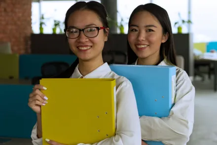Two women smiling and holding binders in a modern office setting.