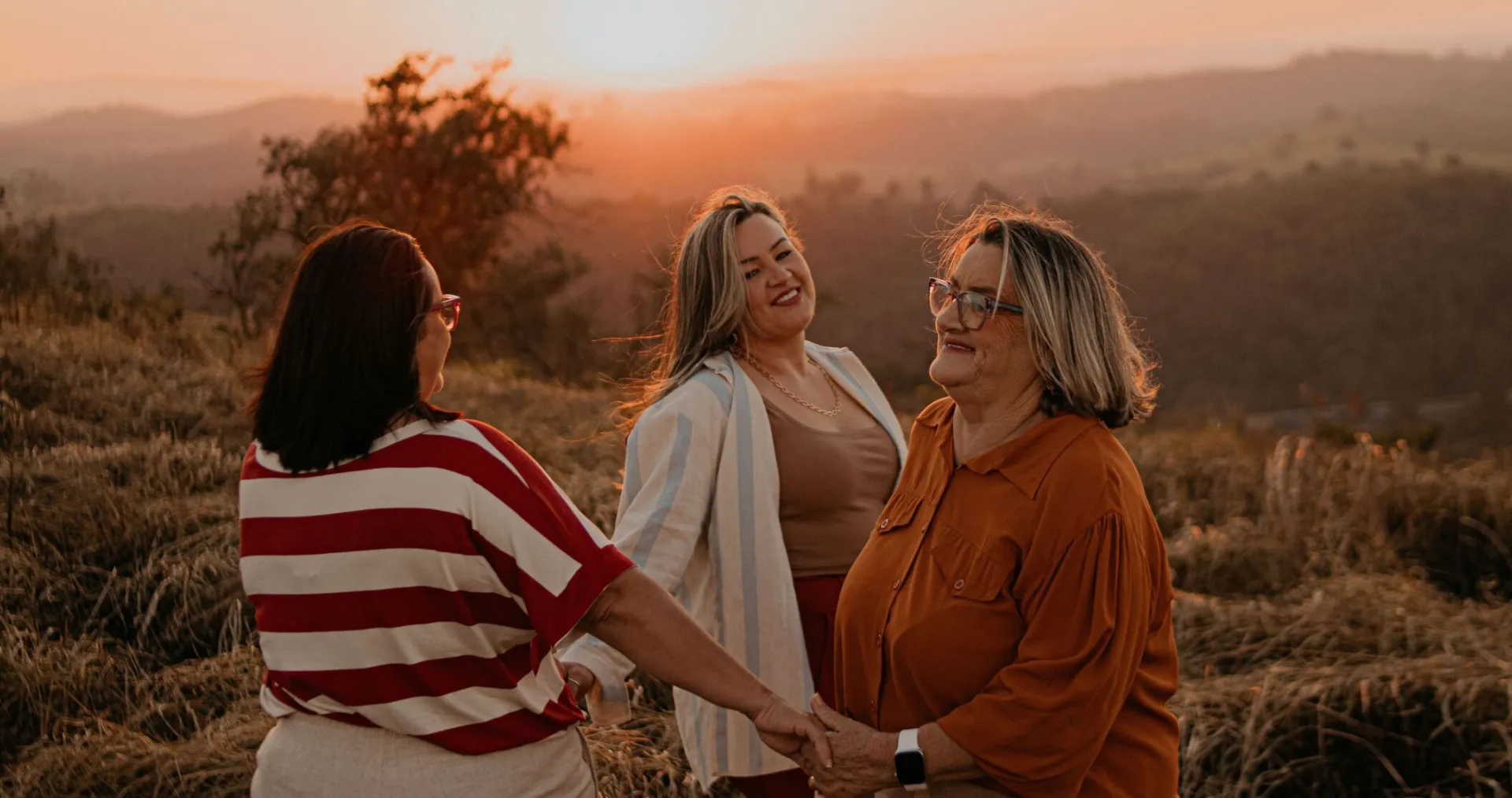Three women enjoying a warm sunset in a São Paulo field, holding hands and smiling.