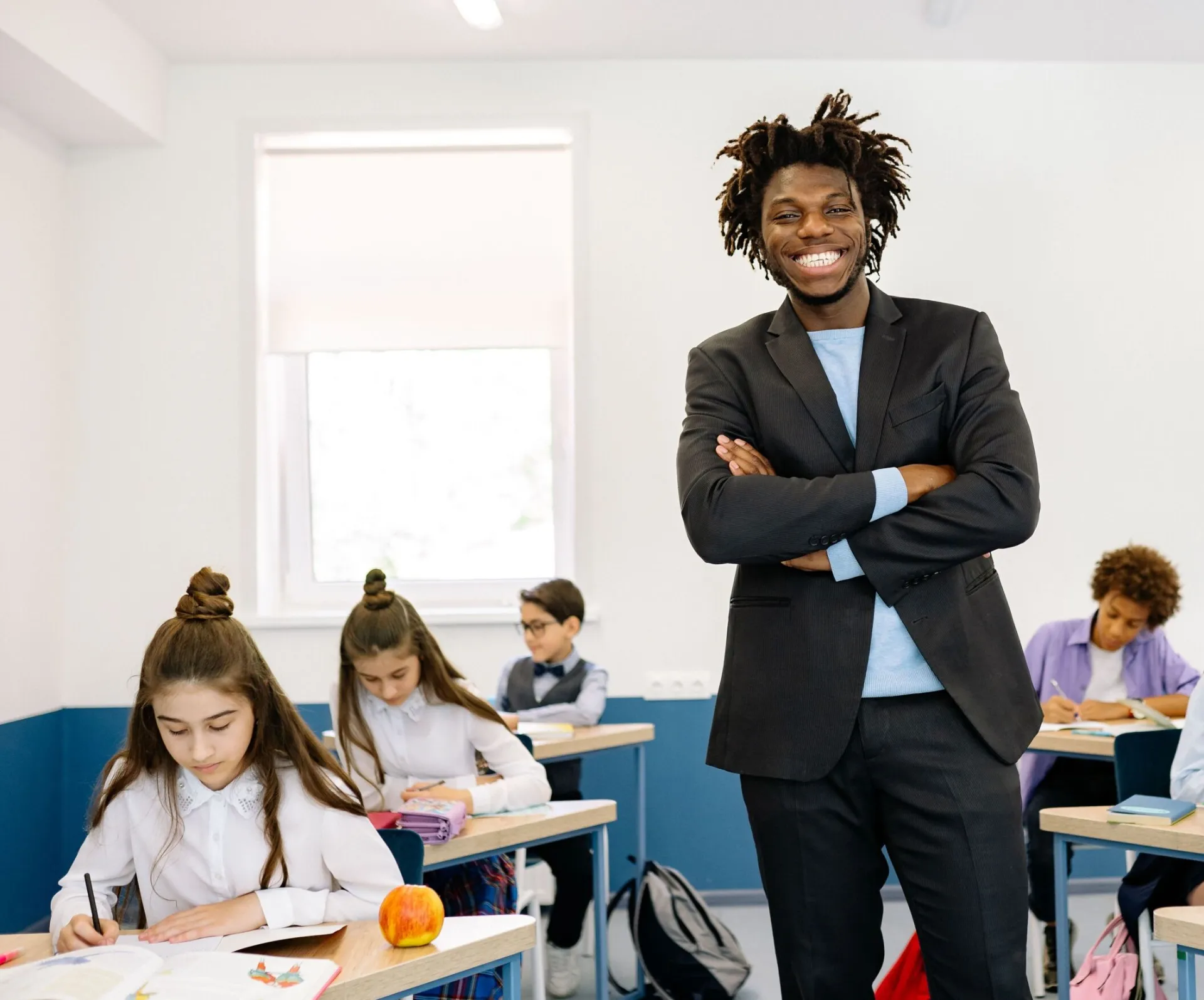 Smiling teacher standing in diverse classroom with students focused on their work.