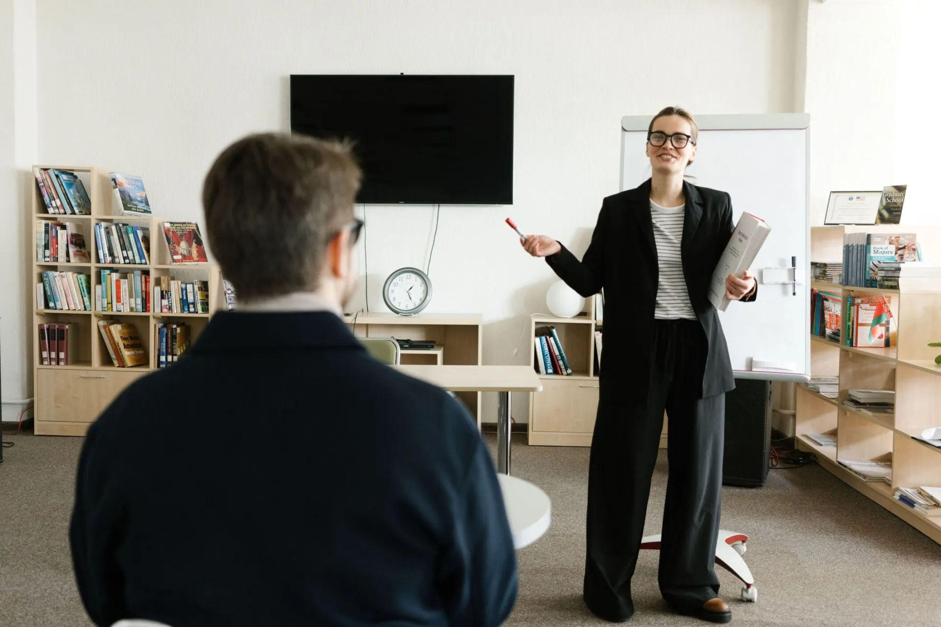 A teacher in a black blazer conducts a session with a student in a library setting.