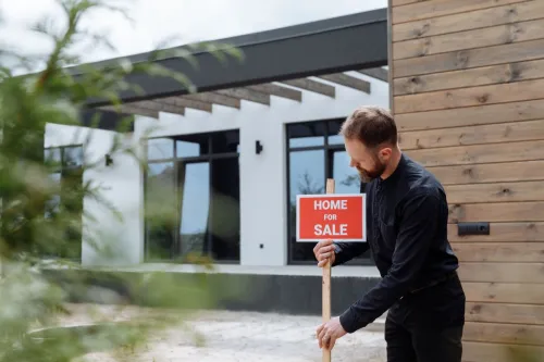 Man placing a home for sale sign outside a modern house for real estate listing.