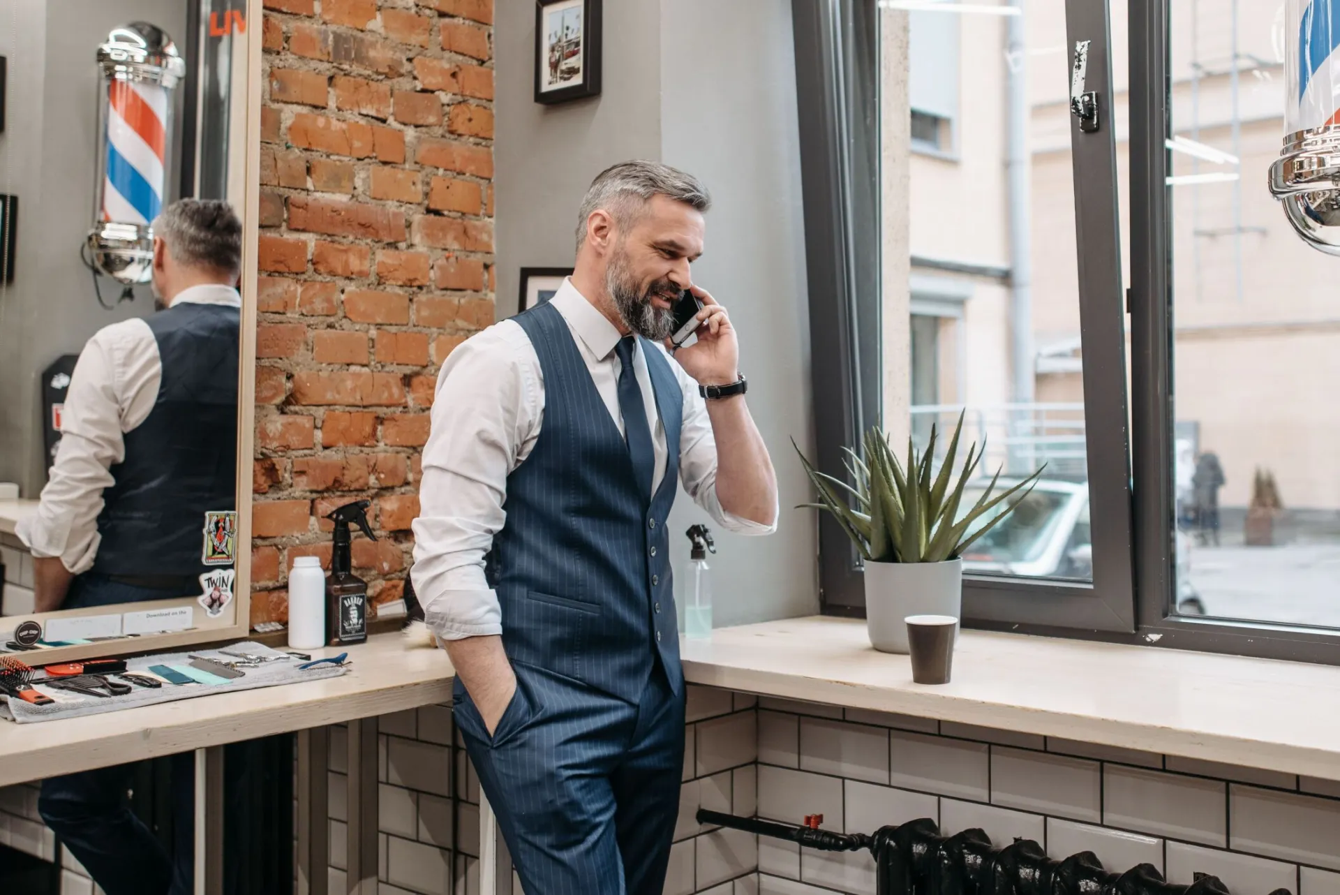 A well-dressed man making a phone call inside a barbershop with modern decor and natural lighting.