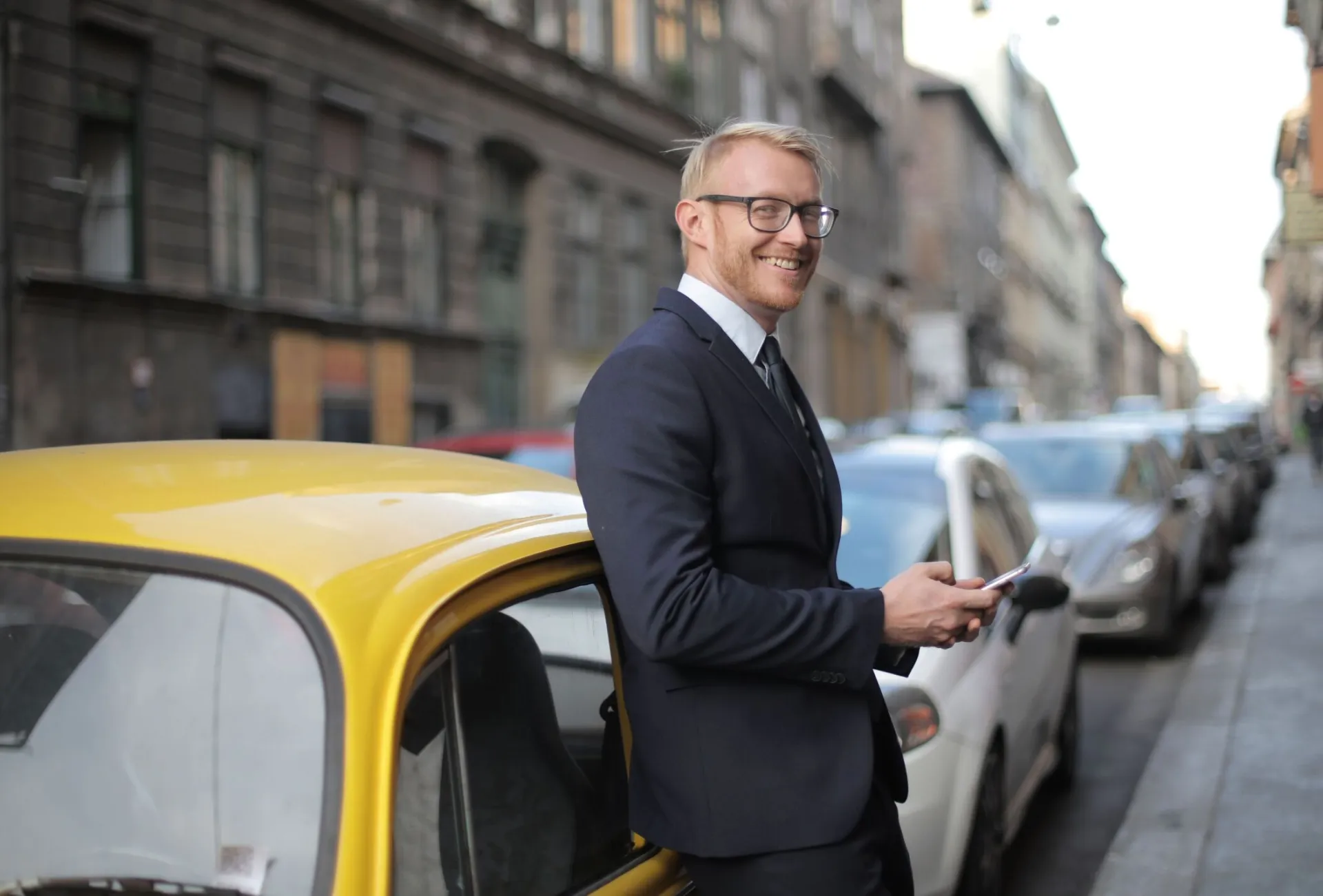 A stylish businessman in formal attire stands confidently by a yellow car on a bustling city street.