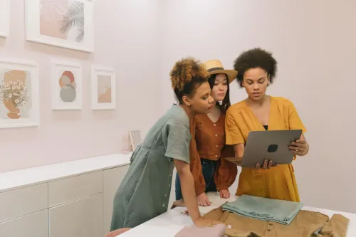 Three diverse women stand indoors, collaborating on a laptop surrounded by modern art.