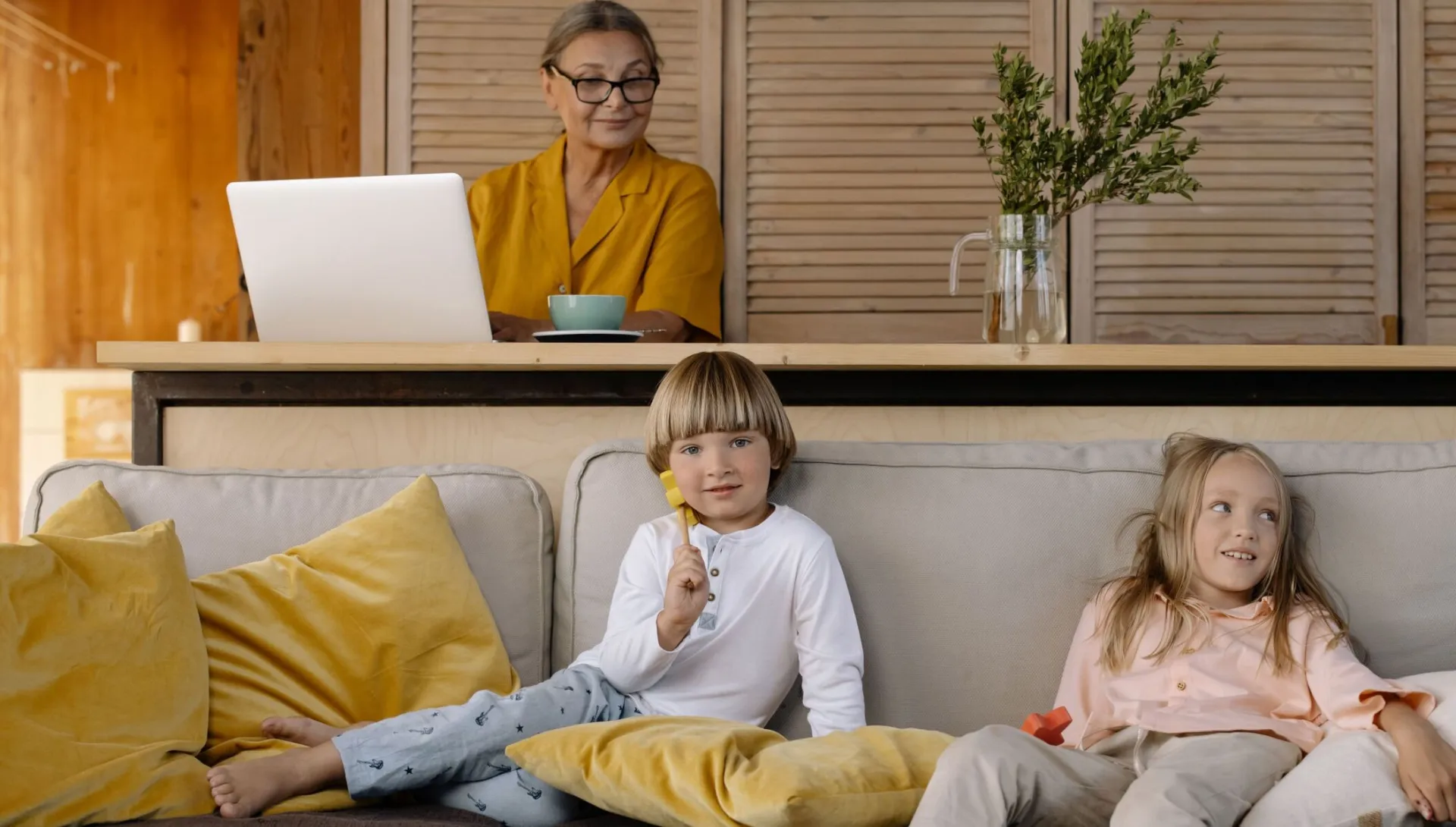 Elderly woman working on a laptop at home while grandchildren relax on the couch.