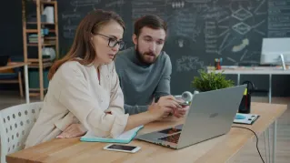 Two young professionals brainstorming at a laptop in a modern office setting.