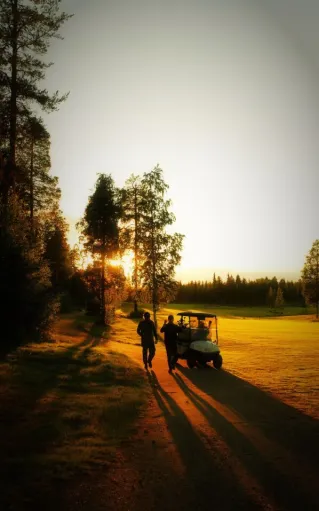 Two golfers walking by a golf cart on a sunny path during sunset in Finland's serene landscape.