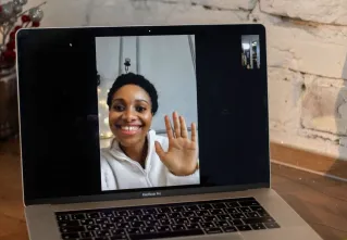 A smiling woman waves during a video call on a laptop, placed on a counter.