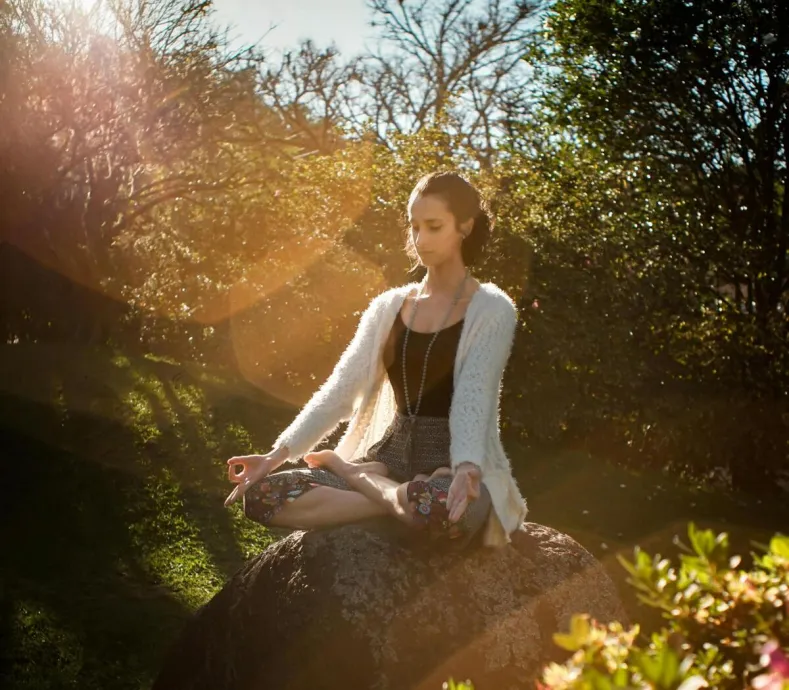 Woman in tranquil meditation pose outdoors, bathed in sunlight, surrounded by lush greenery.